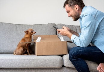 A man and his small Toy Terrier dog unpack a package and take photos of an online purchase.