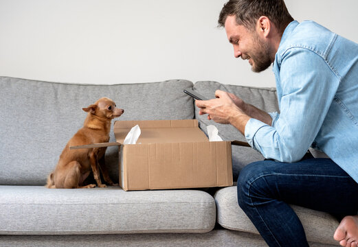 A man and his small Toy Terrier dog unpack a package and take photos of an online purchase.