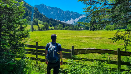 Hiker man walking along lush green alpine meadow with scenic view of Karawanks mountains, Bodental, Carinthia, Austria. Looking at majestic summit of Kosiak. Remote alpine landscape in Austrian Alps - obrazy, fototapety, plakaty