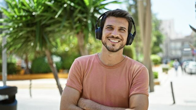 Smiling young man with beard wearing headphones enjoys sunny day in urban park.