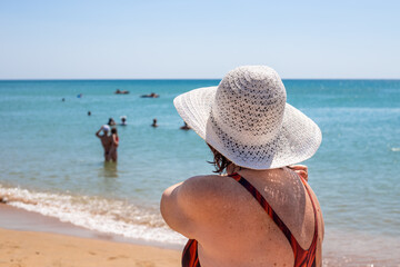 An elderly woman in a hat and swimsuit on the seashore, rear view. Summer holiday on the coast in retirement