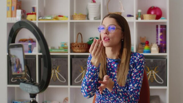 Woman blue floral dress blowing bubble gum front ring light camera setup, colorful shelves background toys books home interior. Female fashion eyewear demonstrates bubblegum playful posture home vlog