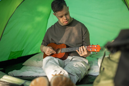 A young man with Down syndrome camping is playing the ukulele