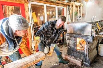 sugar shack, two maple farmers wearing a traditional clothe working doing sugar sirop and puting some wood on fire - 777684471