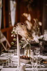 Close-up of a wedding table setting in a warm, illuminated indoor environment with tall candles, a bouquet of dried flowers and eucalyptus in a clear vase, and a basket of bread.