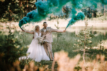 A bride and groom stand in a forest clearing holding blue smoke bombs, with the smoke adding a...