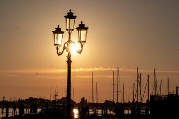 Obraz premium Silhouette of street light and sailing boats in port of charming town of Chioggia at sunrise, nestled in the stunning Venetian Lagoon of the Veneto region in Italy. Tranquil atmosphere in summer
