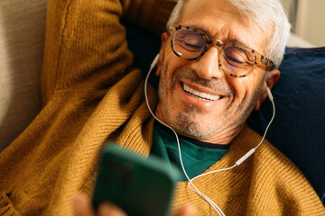 Senior man on couch using his cell phone with earbuds 
