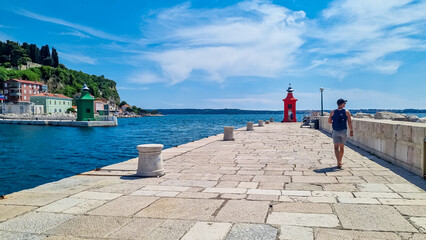 Tourist man walking along picturesque harbor of coastal town Piran. Walking next to the lighthouses overlooking the Adriatic Mediterranean Sea. Summer scene in small port. - obrazy, fototapety, plakaty