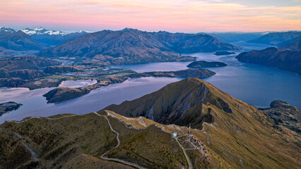 Aerial view of Roys Peak and panoramic backdrop of Lake Wanaka and the Southern Alps of New Zealand during a pink sunrise. 