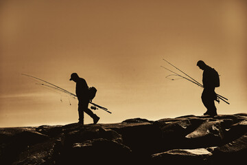 Silhouette of 2 fishermen walking along the stone breakwater at the inlet at Ocean City MD. 2 men with fishing poles and gear head for their favorite fishing spot along the rocks. Atlantic Ocean - obrazy, fototapety, plakaty