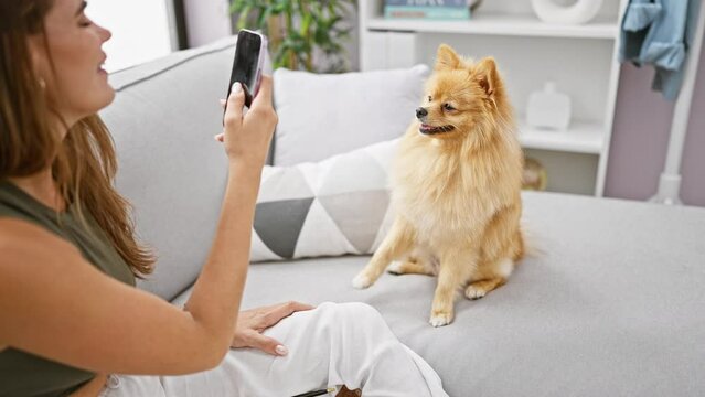 Joyful young hispanic woman sitting on sofa with her beloved dog, capturing a lively portrait on her smartphone, soaking up happiness in her cozy indoor living room at home.