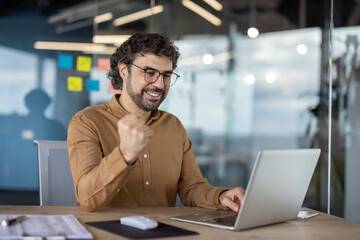 Cheerful businessman with glasses celebrating a success while working on his laptop in a modern office environment.