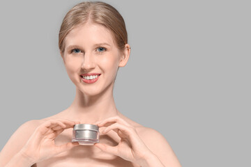 Young blonde woman with jar of facial cream on grey background, closeup