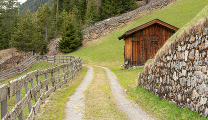 Hiking path between grassy alpine slopes viewing towards pine trees and mountains in Gries,  during early spring time. Last snow on the trail. Besinnungsweg trail in Gries, Längenfeld.Wooden barn.