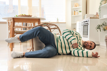 Young man lying on floor after falling from chair in kitchen - obrazy, fototapety, plakaty
