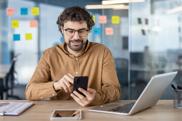 Focused male professional in a casual shirt using smartphone and laptop at a modern workplace with sticky notes in the background.