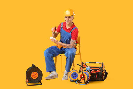 Young male electrician with socket, screwdriver, tool bag and extension cable reels sitting on chair against yellow background