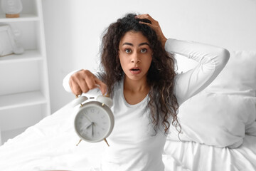 Overslept young African-American woman with alarm clock in bedroom