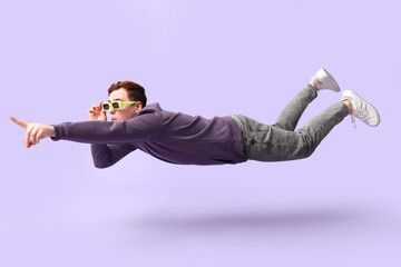 Surprised young man in sunglasses flying and pointing at something on lilac background