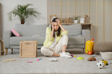 Tired young mother resting among scattered children's toys in living room - obrazy, fototapety, plakaty