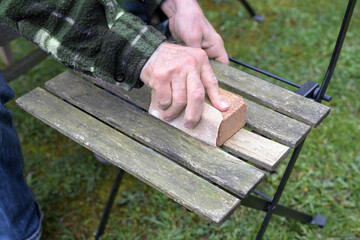 Man restoring a wooden garden chair, sanding and cleaning the weathered wood, removing algae and moss to refresh the outdoor furniture for the next spring and summer season - 777745276