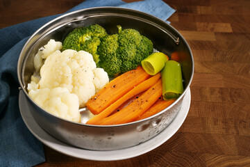 Vegetables such as carrots, broccoli and cauliflower in a metal food steamer basket on a dark rustic wooden table, cooking method to preserve vitamins, minerals and flavor, copy space