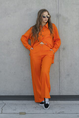 young lady in sunglasses and orange pantsuit at concrete wall