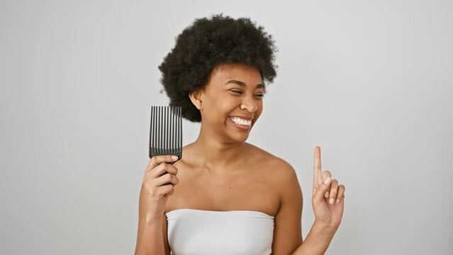 Cheerful african american woman holding comb, wrapped in a towel, smiling and pointing to the side against an isolated white background