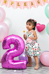 small Latin brunette girl holding a candy in her hands, in the background birthday balloons and a...