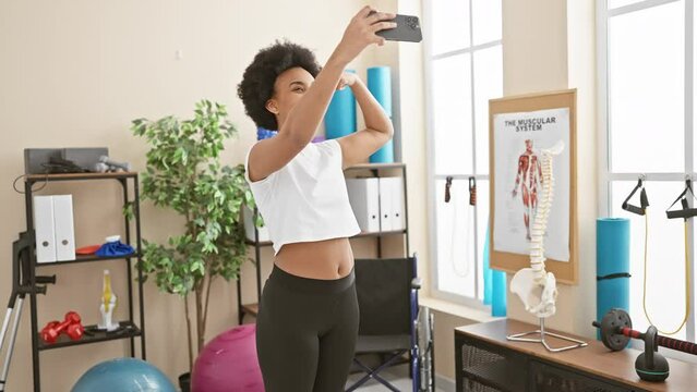 Black woman flexing muscles and taking selfie with smartphone at gym