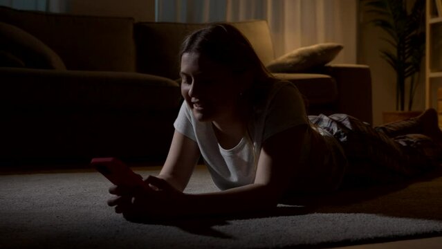 Young woman using phone at night. Adolescent text messaging lying on the floor at home