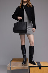 Trendy outfits with black leather bag and black leather boots. Female model with black jacket, luxury clothes and accessories in studio