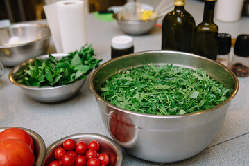 Fresh Arugula Salad Preparation in Stainless Bowls. Stainless steel bowls filled with fresh arugula and tomatoes, accompanied by bottles of olive oil, ready for salad making.