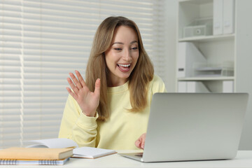 Young woman waving hello during video chat via laptop at white table indoors