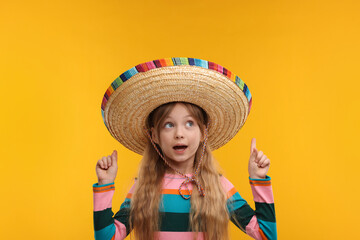 Cute girl in Mexican sombrero hat pointing at something on orange background