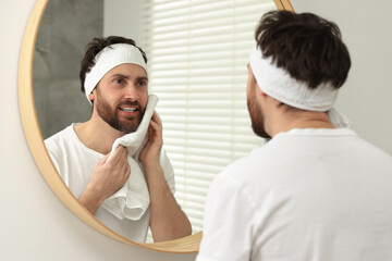 Washing face. Man with headband and towel near mirror in bathroom