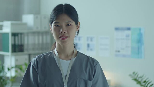 Young Asian female physician wearing scrubs standing in medical office, looking at camera and smiling. Video portrait