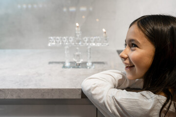 First Candle Hanukkah. Girl Smiles, Admiring Menorah Glow.