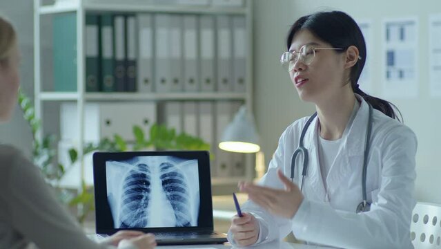 Asian female doctor showing chest x-ray image on laptop screen to patient and explaining diagnosis during checkup in clinic