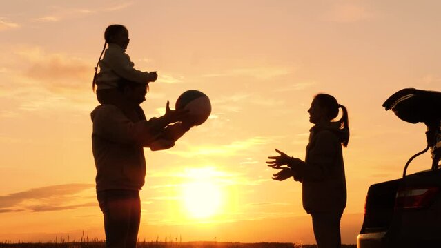 Family playing with ball in nature at sunset, silhouettes. Daughter sits on father's shoulders, throwing ball with mother. Family travel adventures by car, active leisure. Weekend road trip to nature.