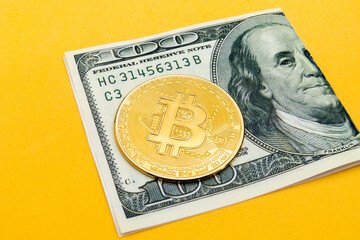 a folded one hundred dollar bill with a bitcoin cryptocurrency on top, focusing on the currency from an inclined and elevated angle on a yellow background, concept of web3 exchange and blockchain