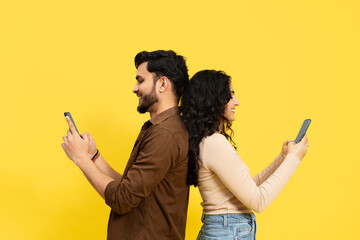Couple Standing Back-To-Back Using Smartphones On Yellow Background