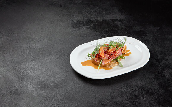 Tuna bruschetta on white plate with sauce on black background. Appetizers with crispy bread and sliced tuna fish. Traditional antipasti - fish bruschetta on dark concrete table. Tuna bruschetta.