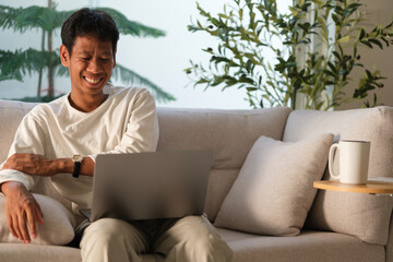 Smiling millennial man using laptop while sitting on comfortable sofa at home.