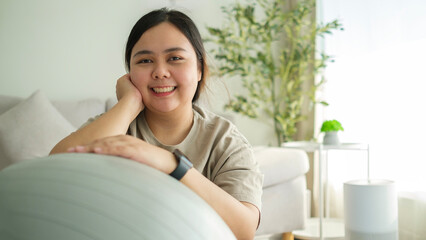 Cheerful overweight young woman  with exercise ball sitting on yoga mat at home. Healthy lifestyle...