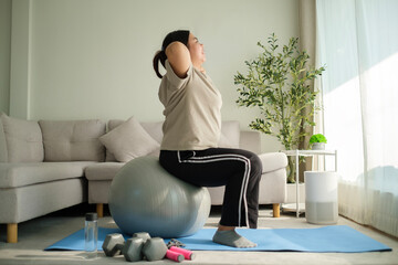 Full length of young overweight woman exercising with fitness ball on mat at home. Healthy...