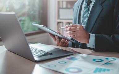 businessperson, executive, graph, lawyer, manager, marketing, search, plan, strategy, analysis. A man is sitting at desk with a laptop and a tablet. He is holding a pen and he is working on a project.
