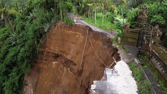 Aerial perspective of the road, destroyed by landslide. Camera tilt down and show sheer drop and cavern. House compound now very close to bluff, with a high risk of future collapse