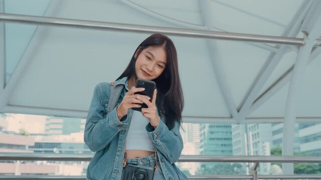 Young asian woman smiling, using mobile smart phone outdoor. Happy beautiful female wearing jeans jacket and holding smartphone at public city. Online technology and communication concept